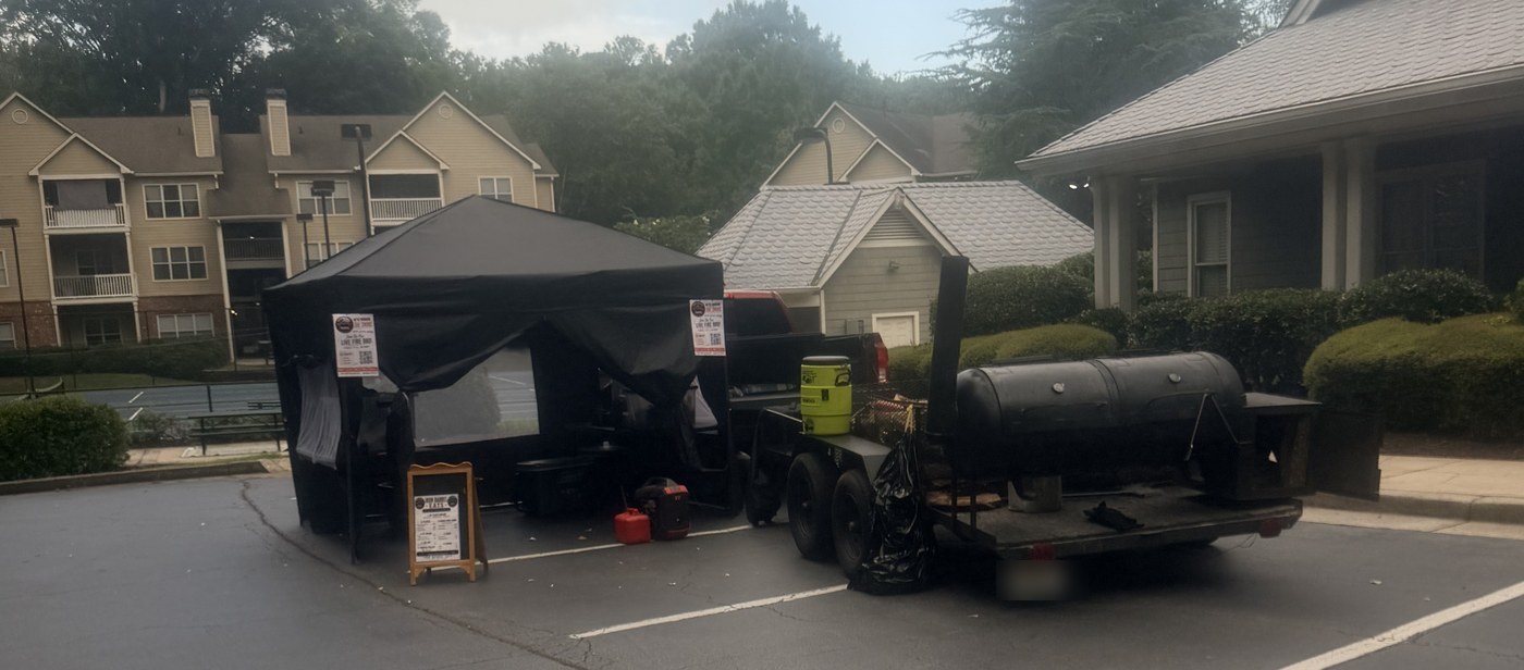 The Iron Barrel Eats tent and offset smoker set up in an apartment community parking lot
